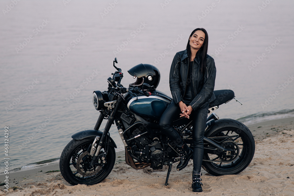 Obraz premium Young woman stands next to motorbike on beach near coast of river.