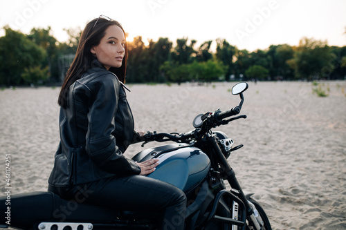 Young woman sits on black motorbike among sandy beach.