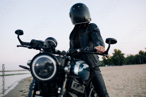 Young woman sits on motorbike in helmet on beach near river.
