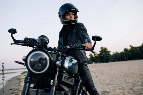 Young woman sits on motorbike in helmet on beach near river.