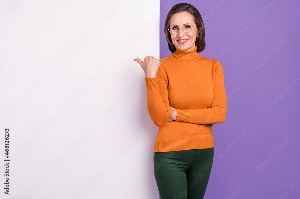 Photo of confident shiny lady pensioner dressed orange turtleneck glasses pointing thumb empty space placard isolated purple color background Photo of confident shiny lady pensioner dressed orange turtleneck glasses pointing thumb empty space placard isolated purple color background
