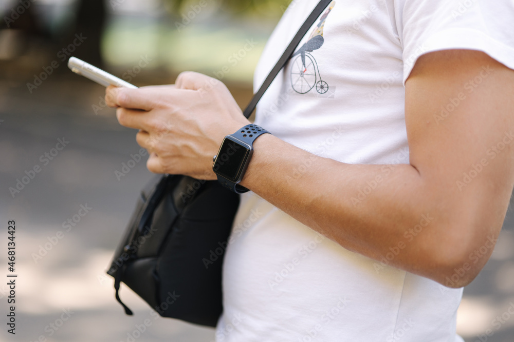 Close-up of man in white t-shirt with black shouler bag using phone outddor. Man has smart watch Close-up of man in white t-shirt with black shouler bag using phone outddor. Man has smart watch