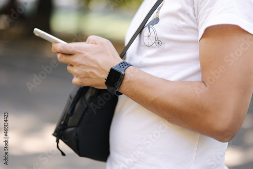 Close-up of man in white t-shirt with black shouler bag using phone outddor. Man has smart watch