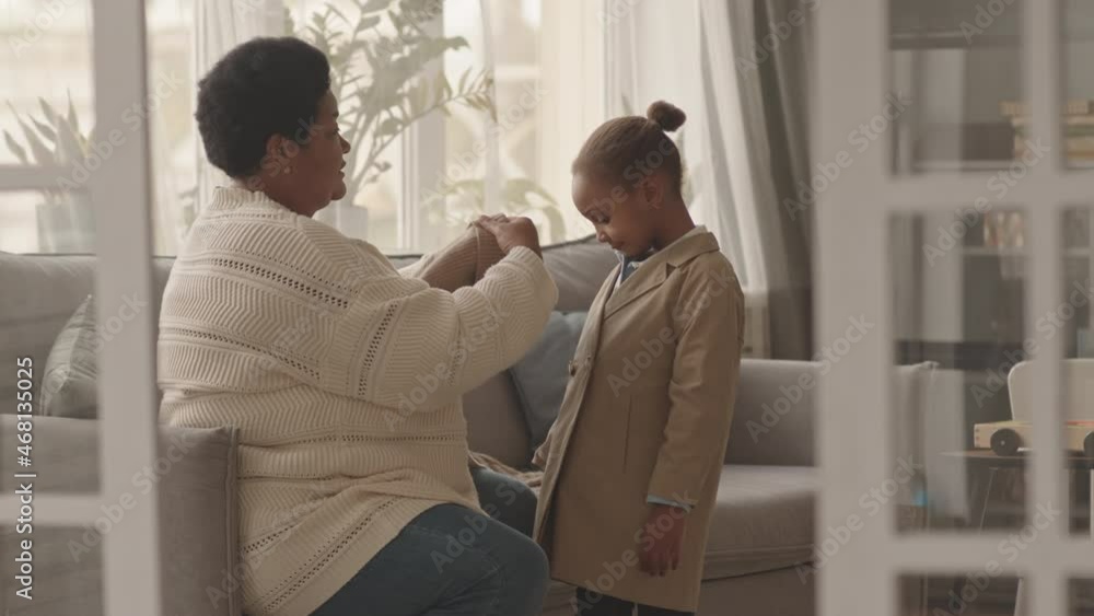 Medium long side view of caring African American senior woman sitting on couch in living room at daytime, dressing up cute little girl in knit hat, scarf, buttoning up trench coat, smiling and talking Medium long side view of caring African American senior woman sitting on couch in living room at daytime, dressing up cute little girl in knit hat, scarf, buttoning up trench coat, smiling and talking