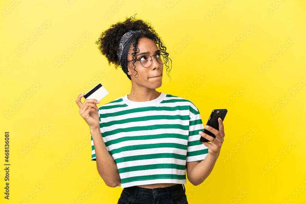 Young african american woman isolated on yellow background buying with the mobile with a credit card while thinking Young african american woman isolated on yellow background buying with the mobile with a credit card while thinking