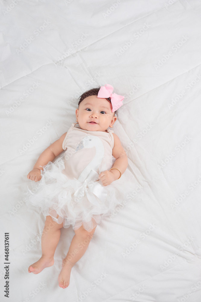 Smiling baby girl toddler in white dress and hair bow decoration lying on bed Smiling baby girl toddler in white dress and hair bow decoration lying on bed