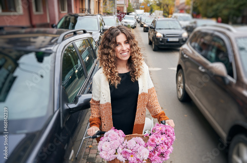 Front view of curly brunette travelling by vintage cycle with big basket of pink flowers. Pretty female wearing brown cardigan riding bike on street between autos. Concept of street style. © anatoliy_gleb