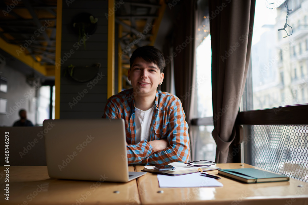 Portrait of smiling male student sitting front open laptop computer while look to the camera. Young man freelancer working remote with online project on netbook Portrait of smiling male student sitting front open laptop computer while look to the camera. Young man freelancer working remote with online project on netbook