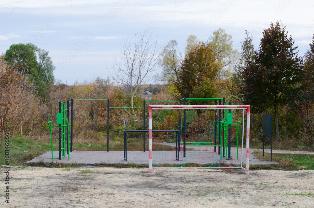 An outdoor sports ground with green and black metal equipment and part of soccer field with football goal in the park with autumn trees. An outdoor sports ground with green and black metal equipment and part of soccer field with football goal in the park with autumn trees.