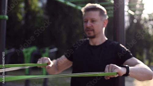 Man guy training with elastic rubber band at the stadium outdoors in black outfit. Athlete male person during workout for arms and chest with additional sport equipmant photo