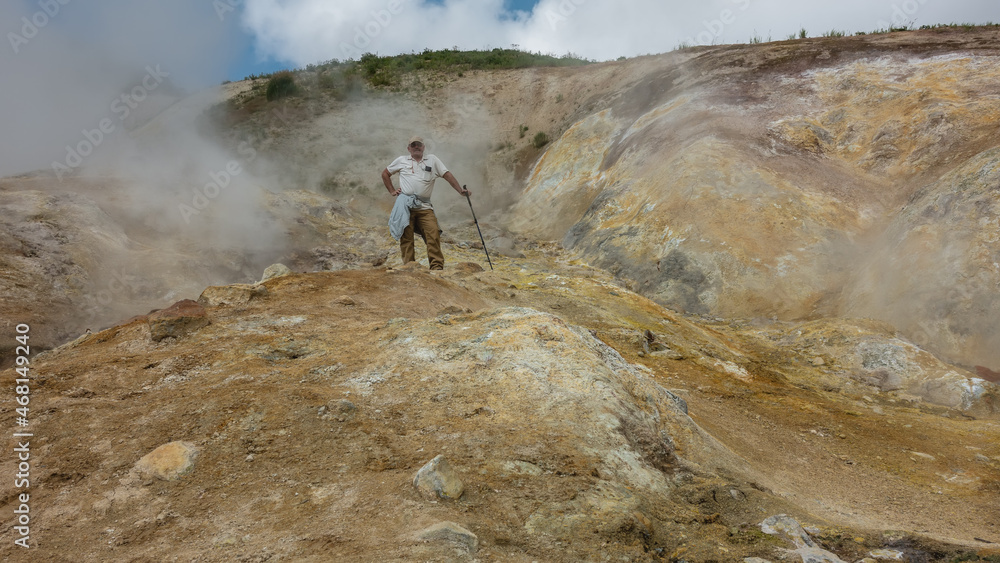 The soil in the geothermal zone is covered with yellow-orange sulfur deposits.  Smoke rises into the blue sky from fumaroles. On the hill there is a male tourist with a trekking pole. Kamchatka