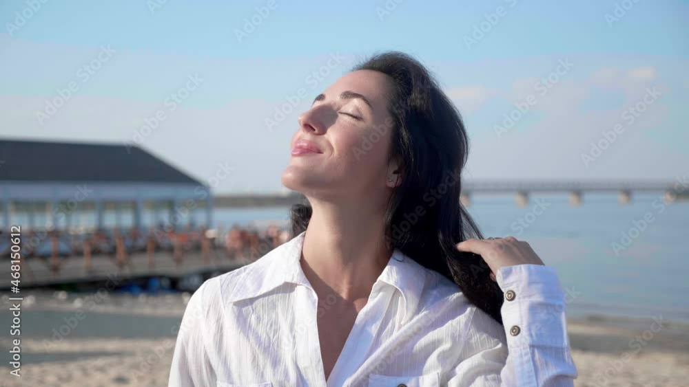 Portrait woman 30 looking at camera standing on the beach with a sea on background. Attractive female in white t-shirt raises eyes. Wind blows airy brown hair shining in sun flares. Portrait woman 30 looking at camera standing on the beach with a sea on background. Attractive female in white t-shirt raises eyes. Wind blows airy brown hair shining in sun flares.