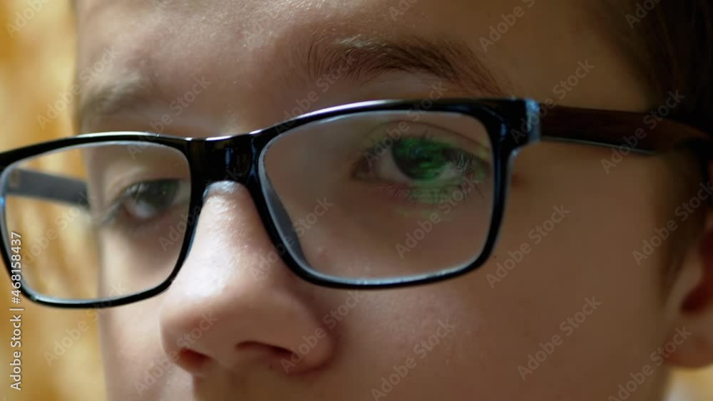 Child with Glasses Watches TV with Display of Running Titles of Screen in Lenses. Close-up of glasses on the eyes of a child with myopia. A teenager looks at the computer monitor, strains vision. Child with Glasses Watches TV with Display of Running Titles of Screen in Lenses. Close-up of glasses on the eyes of a child with myopia. A teenager looks at the computer monitor, strains vision.