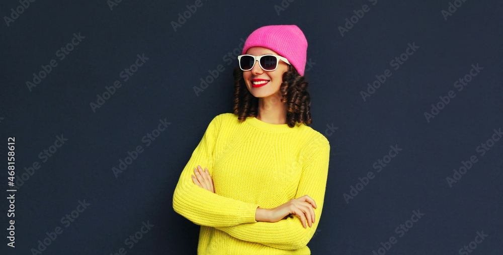 Portrait of stylish smiling young woman wearing an yellow knitted sweater and pink hat on black wall background Portrait of stylish smiling young woman wearing an yellow knitted sweater and pink hat on black wall background