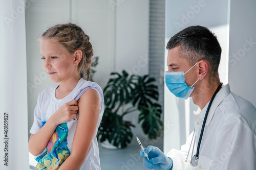 A doctor wearing a medical mask injects a child in the shoulder. Vaccination against coronavirus. COVID-19 vaccine. The doctor vaccinates the kid. A little girl gets a flu shot.