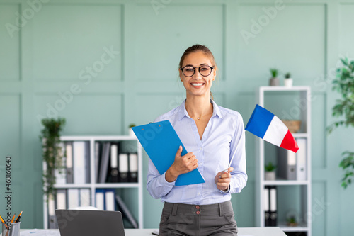 Foreign languages tutoring. Happy teacher holding flag of France and clipboard, standing near workdesk and smiling