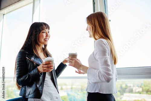 Joyful female colleagues with takeaway caffeine beverage satisfied with friendly communication during coffee break in coworking space, funny Asian and Caucasian woman with disposable cups talking