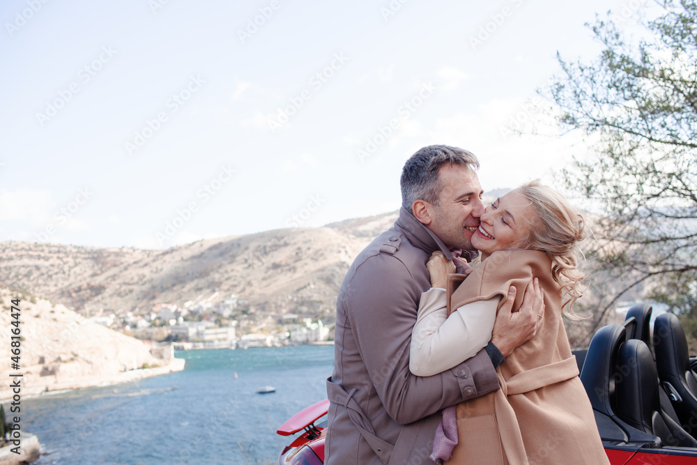 Happy couple, woman and man hugging outdoor and trevelling in the convertible red car. People dressed fashion coat