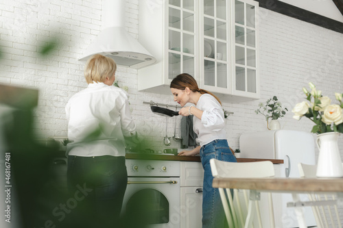 The older mother prepares food in the kitchen, the daughter drinks tea and looks at her mother