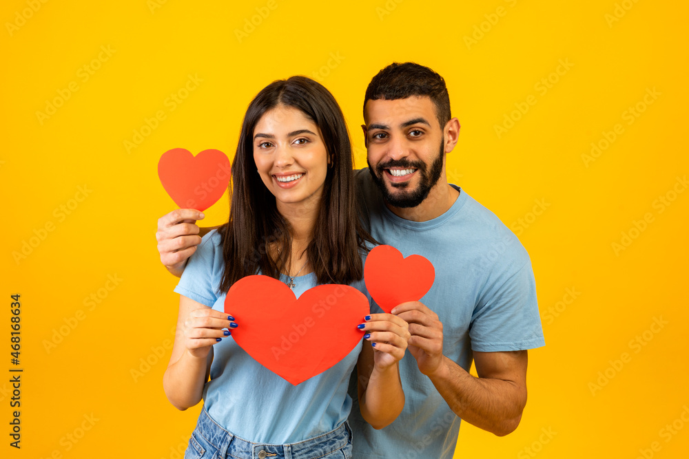 Love is in the air. Romantic arab couple with red paper hearts in hands posing over yellow background, smiling at camera Love is in the air. Romantic arab couple with red paper hearts in hands posing over yellow background, smiling at camera