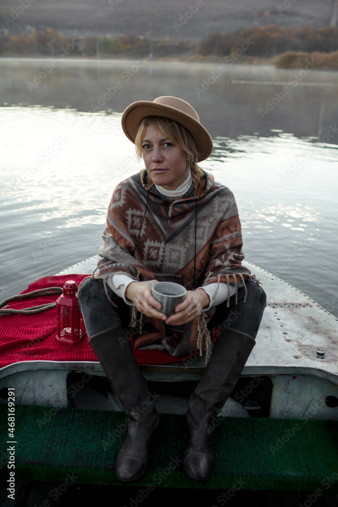 Portrait of woman dressed in poncho, sit in the boat, drink tea and look to the lake, enjoy life, happy in moment Portrait of woman dressed in poncho, sit in the boat, drink tea and look to the lake, enjoy life, happy in moment