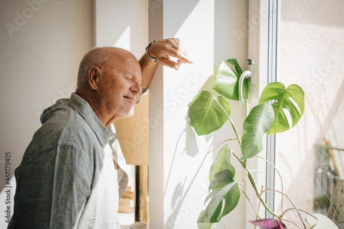 Smiling senior man standing with eyes closed in sunlight by window at home photo