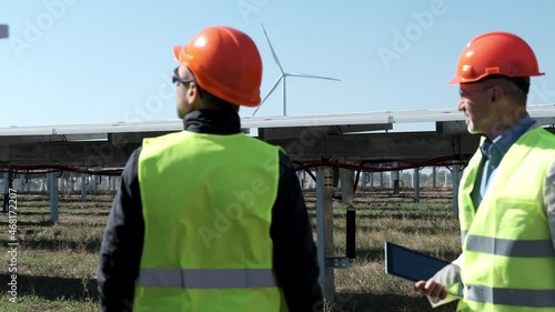 Solar wind energy workers at farm. Mature manager with tablet listens to report of employee near solarcells at construction site backside view photo