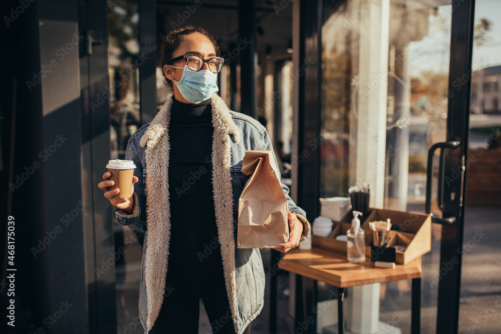 Young woman wearing protective face mask holding takeaway food and coffee to go, standing near restaurant window. Social distancing during quarantine caused pandemic.