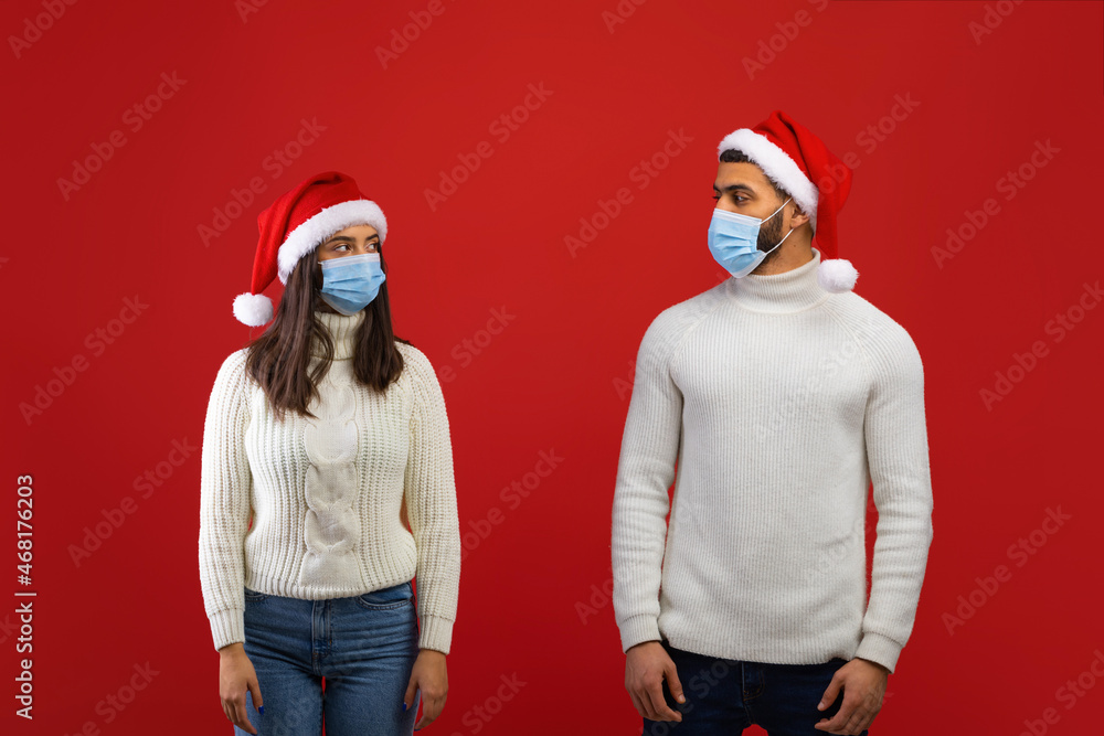 Fototapeta premium Social distancing during covid-19 lockdown. Arab spouses in Santa hats and protective masks looking at each other
