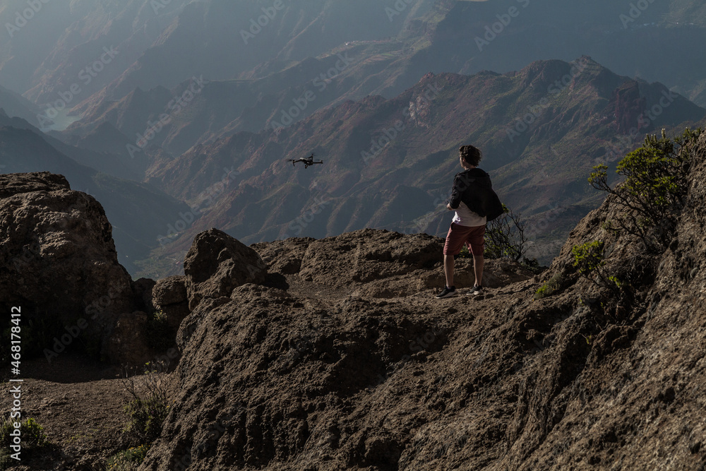 Man piloting a drone on a cliff above a valley with mountains in the background