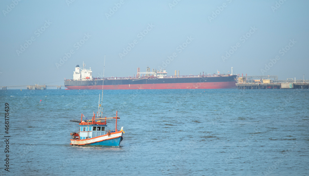 A small blue fishing boat was sailing in the middle of the sea, heading for the shore. There is a sky and a large cargo ship as a backdrop.