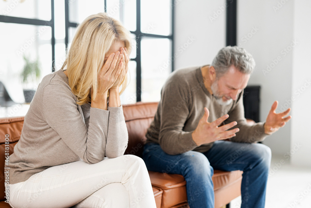 Home abuse concept. Depressed mid-aged woman sits on the couch and crying, an irritated and angry mature man is screaming on the background. A couple is arguing at home