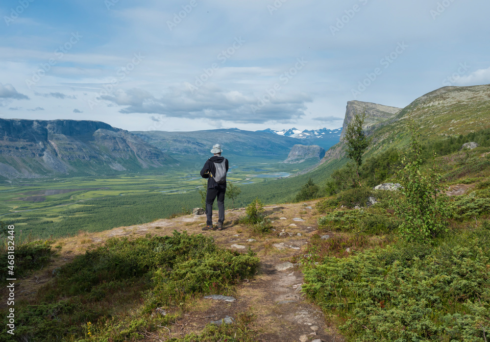 Man hiker admiring view on meandering glacial Rapadalen river delta valley at Sarek national park, Sweden Lapland with big rock, mountains and birch trees. Summer day, white clouds