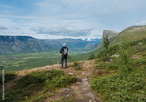 Man hiker admiring view on meandering glacial Rapadalen river delta valley at Sarek national park, Sweden Lapland with big rock, mountains and birch trees. Summer day, white clouds
