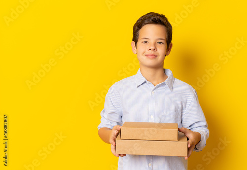 Child boy smiling in blue shirt holding carton boxes isolated over yellow background.