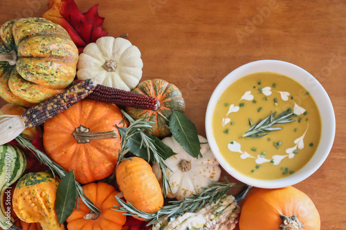 Bowl of butternut squash soup garnished with cream rosemary, and chives, surrounded by various pumpkins, gourds, ornamental corn, and herbs on wood background with copy space.