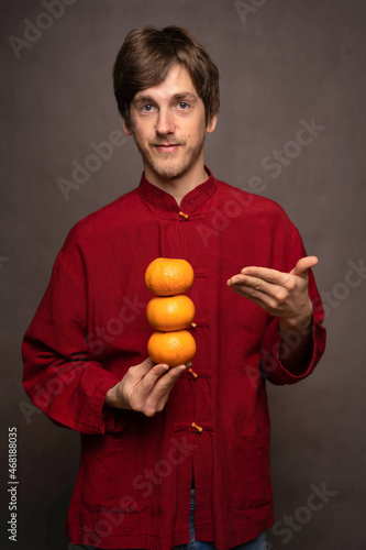 Young handsome tall slim white man with brown hair presentingly holding oranges in red shirt on grey background © PIXbank