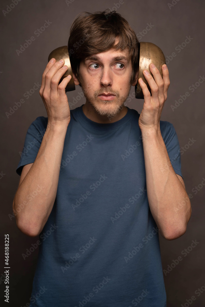 Young handsome tall slim white man with brown hair covering ears with bowls in blue shirt on grey background Young handsome tall slim white man with brown hair covering ears with bowls in blue shirt on grey background