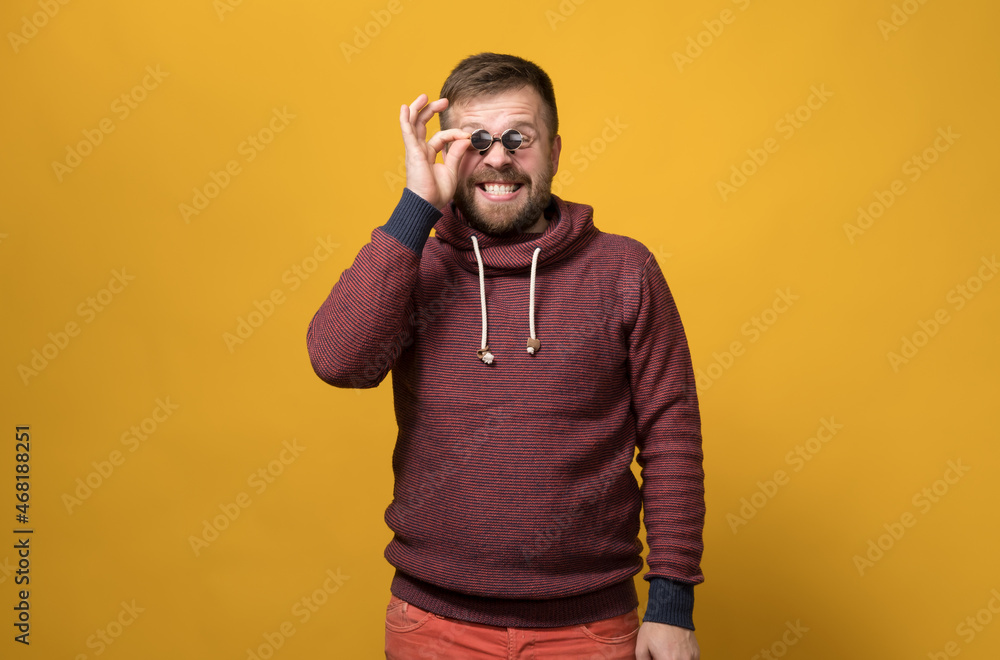 Cheerful Caucasian bearded man holds toy sunglasses like pince-nez to his eyes, with a bizarre grimace and grin. Yellow background. - obrazy, fototapety, plakaty 