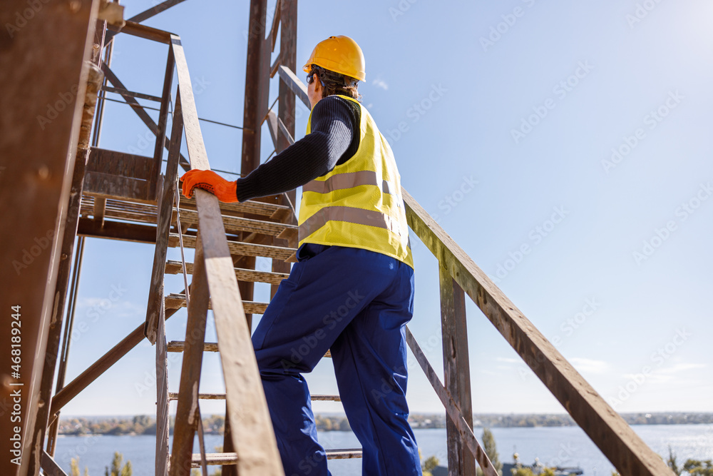Back view of man factory worker wearing work vest and safety helmet while walking up staircase outdoors Back view of man factory worker wearing work vest and safety helmet while walking up staircase outdoors
