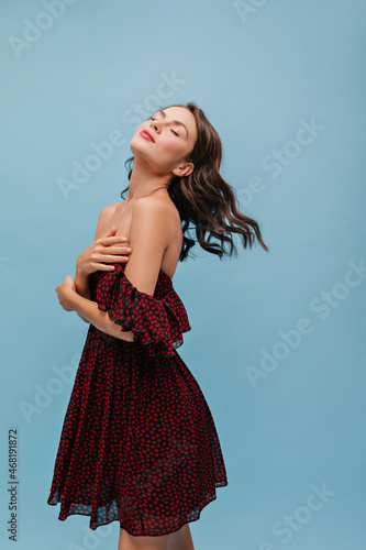 Semi sideways pretty young woman stands on blue background with her head tilted back with closed eyes. Light-skinned lady with bright lipstick is dressed in short summer dress with bare shoulders. photo