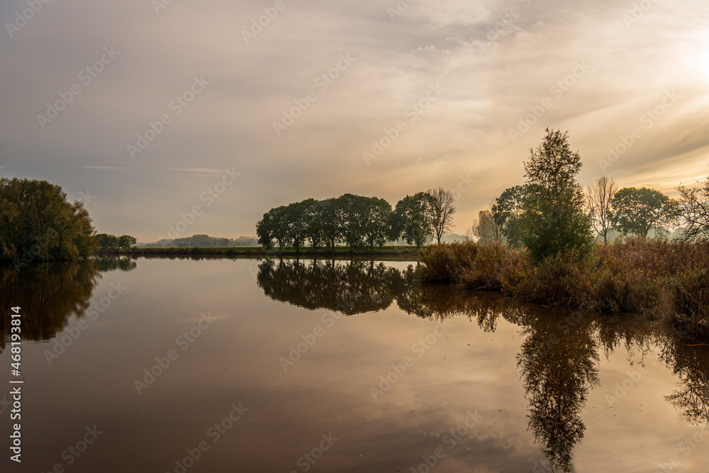 Trees and shrubs reflected in the mirror-smooth water surface of a Dutch lake. It is at the end of a beautiful day in the fall season. The leaves are already changing color.