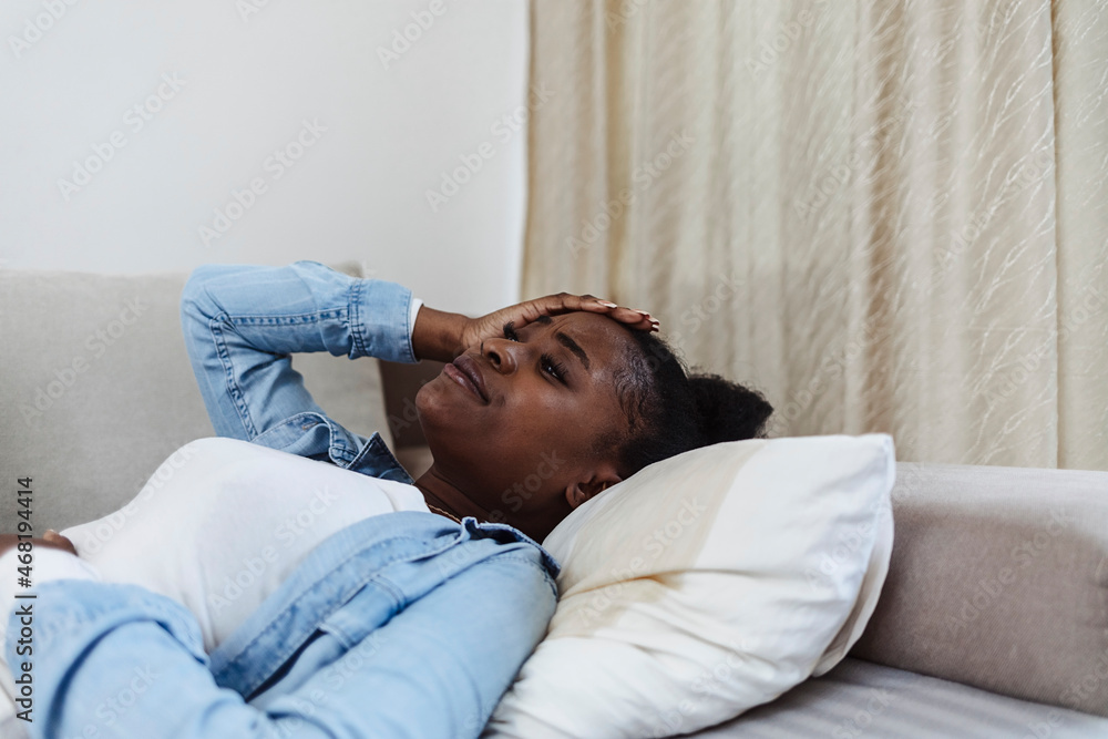 Young African woman with menstrual pain. Black girl having cramps during period, lying on bed. Sad black girl suffering from PMS and menstruation pain, having stomach ache, abdominal pain and headache