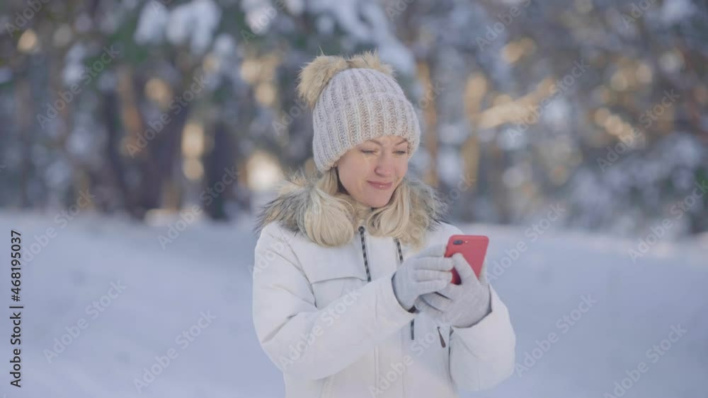 Portrait of cute girl in warm winter clothes talking for video chat using mobile phone and rejoice. Young woman posing outdoors on blurred background of snowy forest. Close up. Slow motion. Portrait of cute girl in warm winter clothes talking for video chat using mobile phone and rejoice. Young woman posing outdoors on blurred background of snowy forest. Close up. Slow motion.