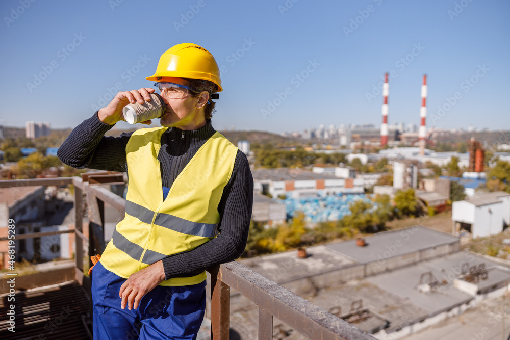 Matured man factory worker wearing work vest and safety helmet while enjoying takeaway hot drink Matured man factory worker wearing work vest and safety helmet while enjoying takeaway hot drink