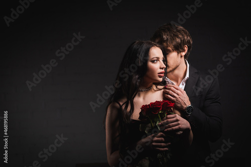 young elegant man hugging sensual brunette woman holding red roses on dark background