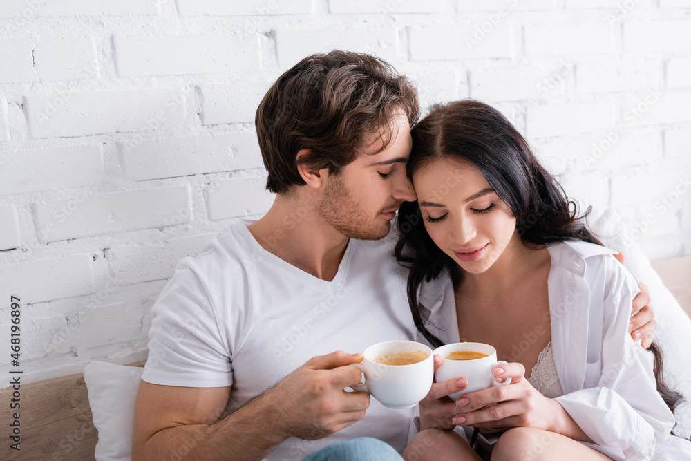 happy young couple sitting in bed with cups of coffee and embracing with closed eyes happy young couple sitting in bed with cups of coffee and embracing with closed eyes