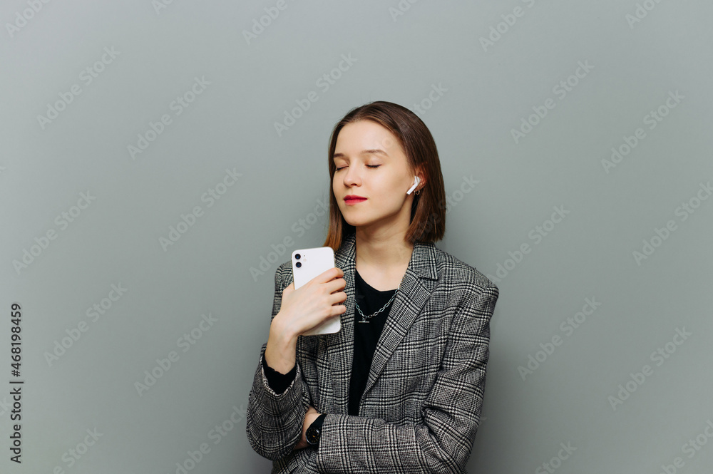 Attractive lady with smartphone in hand and smart casual clothes listens to music in wireless headphones with closed eyes on gray wall background. Attractive lady with smartphone in hand and smart casual clothes listens to music in wireless headphones with closed eyes on gray wall background.