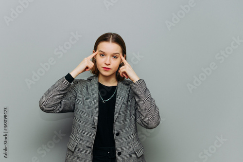 Concentrated woman in a jacket with a serious face stands on a background of a gray wall and looks at the camera and points his fingers at his temple