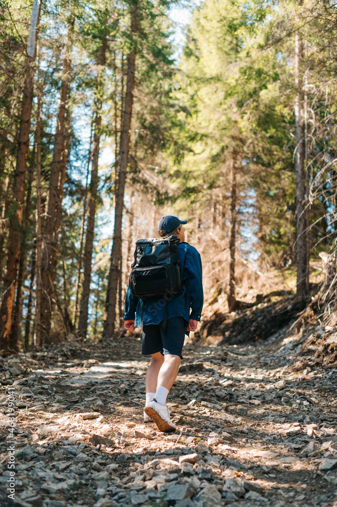 Young hipster man in a shirt and backpack on his back walking on a mountain trail in the woods, rear view. Active rest in the mountains Young hipster man in a shirt and backpack on his back walking on a mountain trail in the woods, rear view. Active rest in the mountains
